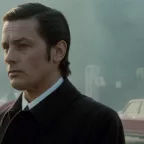Plan rapproché-épaule sur un Alain Delon pensif, dans la rue, tandis qu'une silhouette d'homme s'éloigne de lui au loin, issu du film Big Guns.
