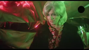 Plan rapproché-poitrine sur l'actrice Chesty Morgan le regard pensif, éclairée par une étrange lumière rose et verte dans le film Supernichons contre mafia.