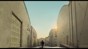 Une silhouette seule, toute petite, vue de dos, marche entre les grand bâtiments de studio de cinéma sous un soleil pesant, dans le film The Fabelmans.