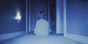 Un petit garçon vu de dos est assis en tailleur au bout d'un couloir, baigné dans un noir et blanc qui vire vers le bleu ; plan du film Skinamarink.