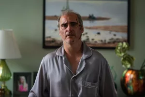 Plan rapproché-taille sur Joaquin Phoenix, fatigué, dans son salon, des portraits de famille en fond, et un tableau représentant une plage sur un mur vert ; issu du film Beau is afraid.