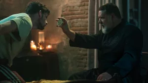 Scène du film L'exorciste du Vatican où le père Amorth montre un médaillon à un jeune en jogging, penché vers lui, dans la chambre de ce dernier seulement éclairé par des bougies.