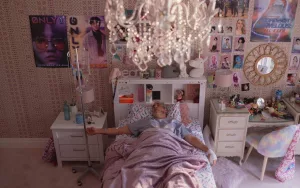 Joaquin Phoenix adulte allongé dans un lit d'adolescent avec des posters aux murs, juste en dessous d'un immense lustre ; il est en tenue d'hôpital et une perfusion est reliée à son bras ; plan issu du film Beau is afraid.