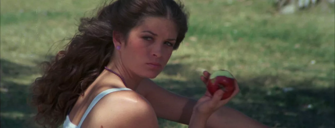 Une adolescente mange une pomme assise dans un parc vert, elle regarde vers nous, dans une moue fermée ; scène du film Sweet sixteen.