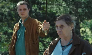 Un jeune homme tient un parapluie au dessus de la tête d'une vielle femme, en forêt, pour la préserver du soleil ; il a une attitude blasée ; plan issu du film Jacky Caillou de Lucas Delangle.