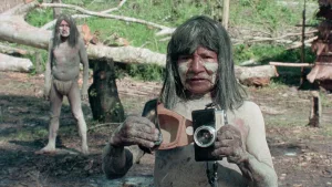 Un indigène d'Amazonie tient un appareil photo dans sa main, au niveau de sa poitrine, l'air un peu dépassé, en pleine jungle, dans le film Cannibal Ferox.