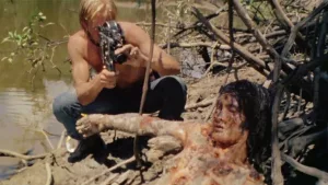 Un journaliste torse nu et en jean filme avec une caméra Super 8 un corps d'indigène en train de se décomposer au bord du fleuve Amazone ; scène du film Cannibal Holocaust de Ruggero Deodato.
