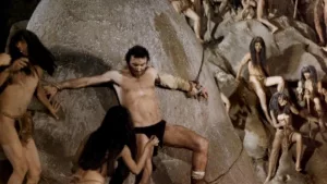 Massimo Foschi, attaché à une paroi rocheuse par des cordes, en slip, tente de s'écarter d'une indigène qui l'approche dans le film Le dernier monde cannibale de Ruggero Deodato.