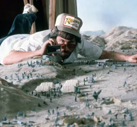 Steven Spielberg sur le tournage d'Indiana Jones, allongé sur une maquette de plaine avec des petits soldats verts, un objectif rivé à l’œil.