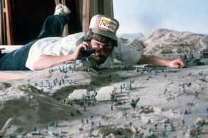 Steven Spielberg sur le tournage d'Indiana Jones, allongé sur une maquette de plaine avec des petits soldats verts, un objectif rivé à l’œil.