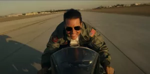 Tom Cruise vu de face tout sourire dans le film Top Gun : Maverick roulant sur une large route en moto grosse cylindrée.