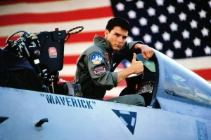 Tom Cruise jeune pilote dans le premier Top Gun, est assis dans le cockpit et se tourne vers l'objectif en faisant un pouce ; en fond, le drapeau américain.