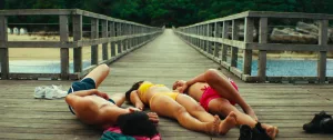 Sur la jetée en bois d'un lac, trois corps, deux jeunes hommes et une jeune femme, sont allongés pour bronze ; on ne voit pas leur visage ; plan issu d'un film de Simon Rieth.