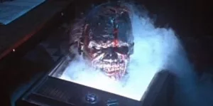 Une tête de mort-vivant ensanglanté et avec un sourire diabolique surgit d'un écran de télévision brisé, dans de la fumée ; plan issu du film The Video Dead.
