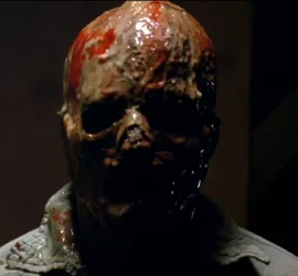 Plan rapproché-épaule en contre-jour sur un homme dont la peau a fondu, ne reste que son crâne et un peu de sang et de chair marron, coulante ; issu du film Le monstre venu de l'espace.