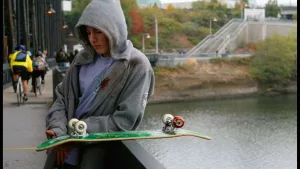Un adolescent capuche relevé attend mélancoliquement sur la rambarde d'un pont, son skateboard près de lui, dans le film Paranoid Park.