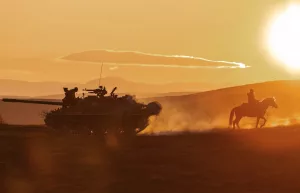Sous un soleil de crépuscule qui se couche sur des dunes faisant penser à un désert, un tank suit un homme à cheval ; plan du film Sisu : de l'or et du sang.