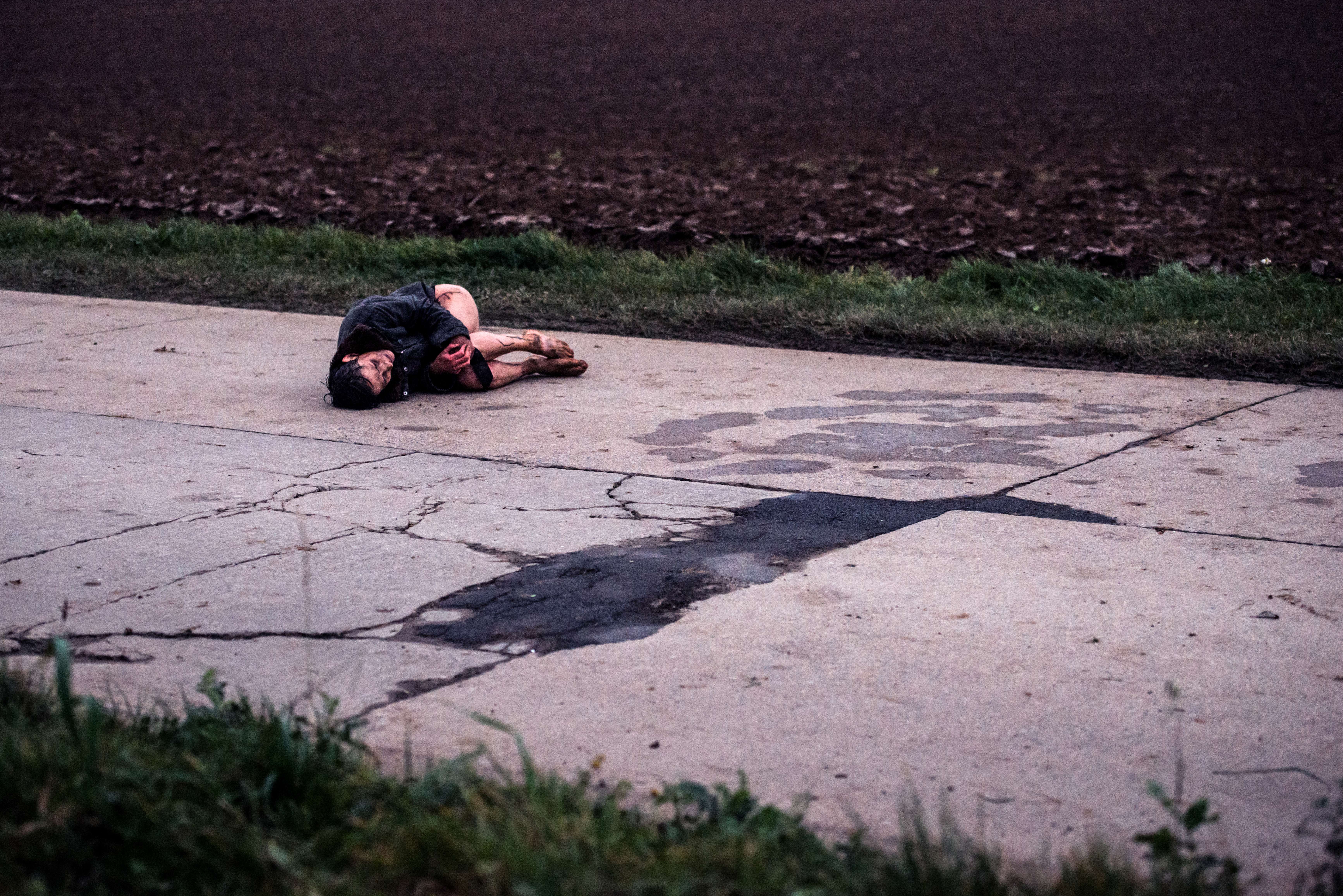 Un homme blessé et sans pantalon est recroquevillé sur un trottoir délabré, en pleine rue ; plan issu du film Megalomaniac.