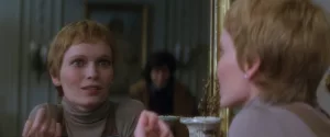 Mia Farrow regarde le reflet de son interlocuteur dans le miroir, d'un air proche de la folie, avec un étrange sourire absent, dans le film Le cercle infernal.