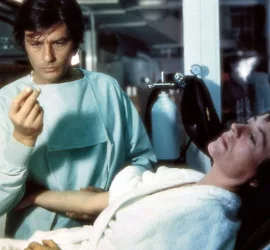 Alain Delon en tenue de médecin observe d'un air intrigué une gélule qu'il tient entre ses doigts; sur le lit d'hôpital tout près de lui en, Annie Girardot prête à une injection, dans une pose abandonnée ; scène du film Traitement de choc.