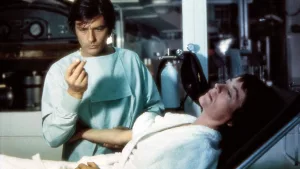 Alain Delon en tenue de médecin observe d'un air intrigué une gélule qu'il tient entre ses doigts; sur le lit d'hôpital tout près de lui en, Annie Girardot prête à une injection, dans une pose abandonnée ; scène du film Traitement de choc.