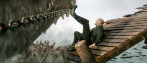 Sur une jetée en bois, Jason Statham repousse du pied un mégalodon gueule ouverte dans le film En eaux très troubles.