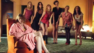 Laura Dern, au premier plan, est assise sur un fauteuil en peignoir rose, boudeuse, dans un salon aux murs jaunes et à la moquette verte ; derrière elle cinq jeunes femmes prennent une pose enfantine ; plan issu du film Inland Empire.