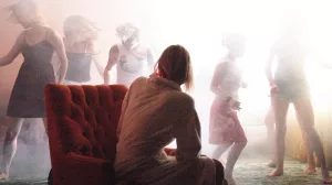 Laura Dern vue de dos assise sur son fauteuil observe cinq jeunes femmes danser dans une brume très illuminé par le soleil à l'extérieur ; scène dans le salon du film Inland Empire.