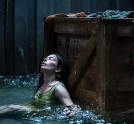 Une femme qui semble épuisée, est assise, adossée à une vieille et massive caisse en bois ; de l'eau arrive à sa poitrine ; plan issu du film Nowhere.