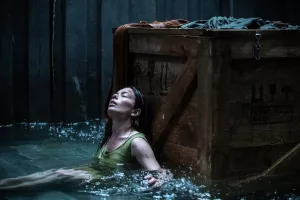 Une femme qui semble épuisée, est assise, adossée à une vieux et massif cagot en bois ; de l'eau arrive à sa poitrine ; plan issu du film Nowhere.
