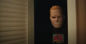 Un tueur portant un masque d'homme blondinet et souriant se terre près d'une fenêtre, aux volets baissés, dans le film Totally Killer.