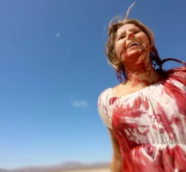 Une jeune femme hilare marche dans le désert portant une robe blanche ensanglantée dans le film The Outwaters.