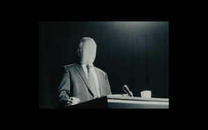 Un homme en costume-cravate est à un pupitre, avec un sac en toile sur la tête ; scène du film From Beyond pour notre carnet de bord 2 Court-Métrange.