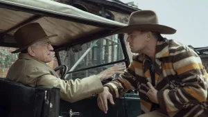 Robert de Niro, au volant de sa vieille voiture, tend sa main vers le bras de Leonardo Di Caprio, penché sur lui, qui l'écoute attentivement, dans le film Killers of the flower moon.