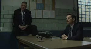 Les deux policiers de la série Manhunter, réalisée par David Fincher, en salle d'interrogatoire ; l'un est assis à une table, avec devant lui un micro ; l'autre est assis contre le mur, en hauteur, sur sa droite.