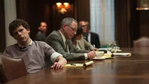 Jesse Eisenberg dans le film The Social Network de David Fincher ; il est à une réunion qui semble profondément l'ennuyer, se tenant de manière désinvolte tandis que des personnes plus âgées, en costume cravate, prennent des notes à côté de lui.
