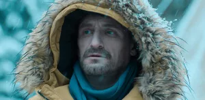 Plan rapproché-épaule sur le visage pensif de Jean-Paul Rouve, dans un manteau d'hiver à capuche jaune, sous la neige ; issu de Polar Park.