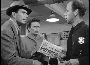 Jim Austin et son épouse face à un policier dans un commissariat, alertes et en sueur, tenant un journal dans les mains, dans le film La ville captive.