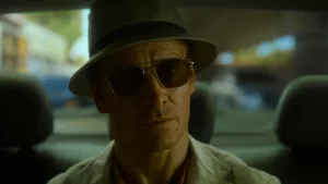 Plan rapproché-épaule sur The Killer, avec lunettes de soleil et chapeau, à l'arrière d'un taxi.