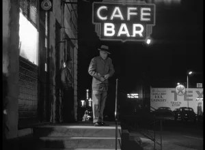 Un homme descend un petit escalier, de nuit, sous une enseigne Café Bar dans le film noir La ville captive.