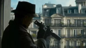 Le tueur du film The Killer observe par la fenêtre avec une longue vue l'immeuble haussmannien, sous un ciel bleu, d'en face. L'homme est vu en contre-jour, de trois-quart, et il porte un long manteau et un chapeau.