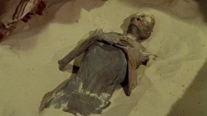 Une momie calcifiée repose dans du sable, presqu'ensevelie, dans le film de la Hammer : Dans les griffes de la momie.
