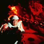 Un homme lutte pour ne pas tomber dans une grotte illuminée de rouge, comme si de la lave arpentait les murs, dans le film Zu les guerriers de la montagne magique.