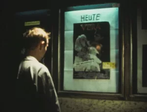 L'acteur Daktari Lorenz, vu de dos, regarde la tête baissée l'affiche d'un faux film d'horreur appelé Vera sur les murs d'un cinéma, de nuit ; scène du film Nekromantik.