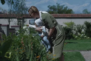Une jeune femme allemande blonde fait découvrir des fleurs à son nourrisson, qu'elle penche vers le tiges pour qu'il puisse les toucher, dans le jardin de la maison du film The Zone of Interest.