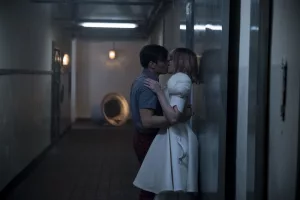 Dans le couloir d'un immeuble, un homme et une femme s'embrassent, adossés contre un mur ; tout au fond du couloir, une large corbeille renversée ; scène du film Halfway home.