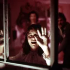 La petite Audrey Rose effrayée tente de s'échapper de sa chambre plongée dans une lumière rose, en criant et posant ses mains sur la vitre ; derrière elle ses parents s’apprêtent à l'en empêcher.