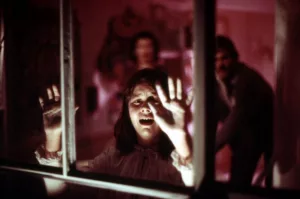 La petite Audrey Rose effrayée tente de s'échapper de sa chambre plongée dans une lumière rose, en criant et posant ses mains sur la vitre ; derrière elle ses parents s’apprêtent à l'en empêcher.