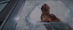 En contre-plongée, une vitre brisée ; Bruce Willis se tient dans le trou dans le verre, droit, toisant l'adversaire qui, on le devine, vient de tomber de l'immeuble ; plan issu du film Die Hard : Piège de Cristal.