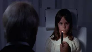 Un vieil homme, vu de dos, tient une bougie sous le regard fixé sur la flamme de la petite fille nommée Audrey Rose, dans le film de Robert Wise.